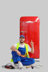Happy male construction worker with professional tools and battery tester pointing at red fridge on...