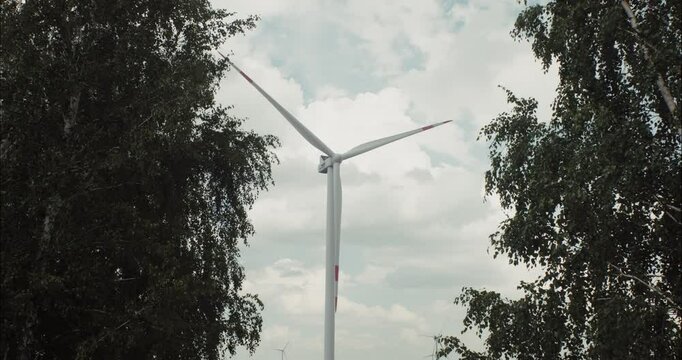 Wind turbine rotating between green trees under cloudy sky
