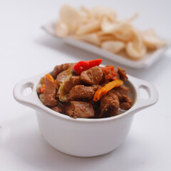 Close-up of a spicy chicken stir-fry in a white bowl with chili peppers and crackers in the background on a white surface