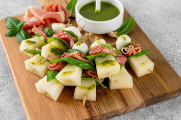 Appetizer with melon, mozzarella, prosciutto ham and fresh basil on stick with pesto sauce on a stone background. Finger food. Summer salad.
