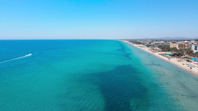An aerial panorama showcasing the beautiful beachfront of Hammamet,lined with lush palm trees and vibrant resorts.A parasail adds a touch of excitement as it glides over the tranquil turquoise waters.