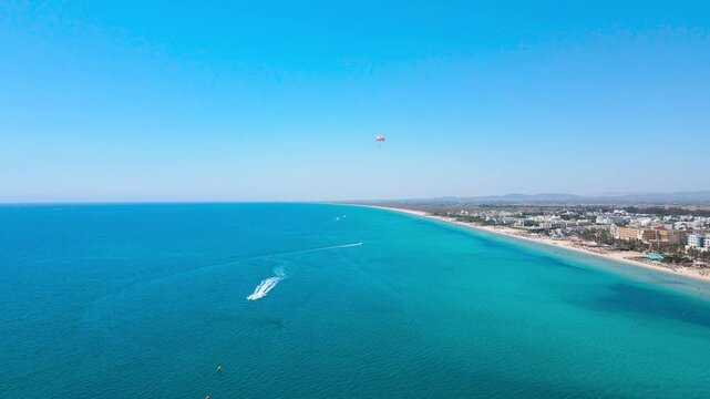An expansive aerial shot capturing the stunning coastline of Hammamet, Tunisia. The crystal-clear turquoise waters stretch to the horizon, with a parasail gracefully soaring above.