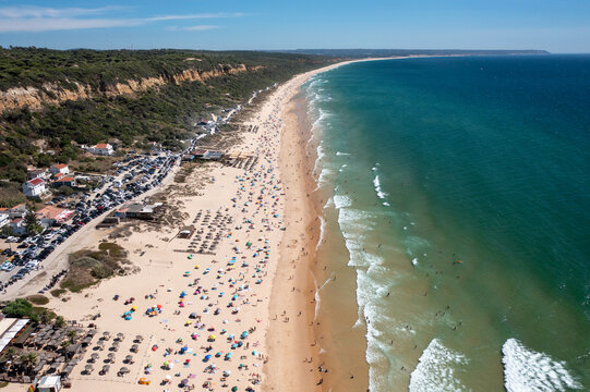 Aerial drone shot of Praia de Fonte da Telha beach in Costa de Caparica, Portugal. Sunbathers and swimmers enjoy the packed beach while the parking lot fills