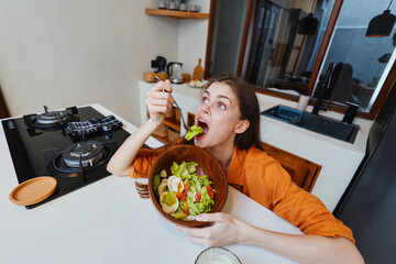A young woman excitedly enjoying a fresh salad in a modern kitchen, showcasing healthy eating...