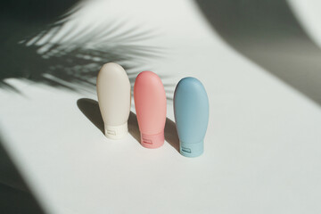 Mock up with travel size cosmetic bottles of pink, blue and white colors for shampoo, conditioner and body care lotion for vacation or journey with palm leaves shadows on background 