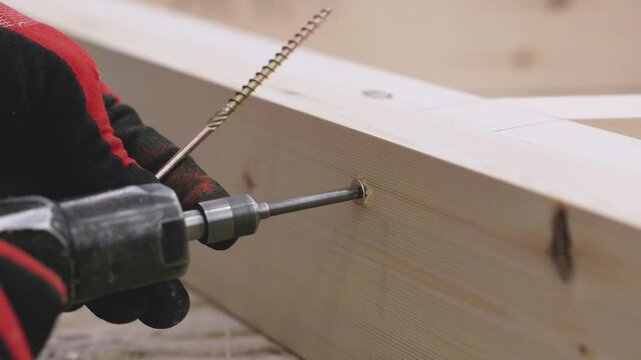 A carpenter uses a power drill to insert a screw into a wooden joint, aligning the pieces precisely. Gloved hand holds the timber, ensuring steady placement and accuracy