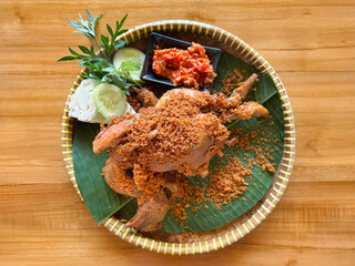 Traditional Indonesian marinated fried chicken with local chili sauce and herbs crackers. Served on a round basket with banana leaf, on a wooden table.