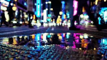 Vibrant city nightlife video with a low-angle shot, capturing neon lights reflecting in a puddle, creating a dynamic and colorful urban scene.