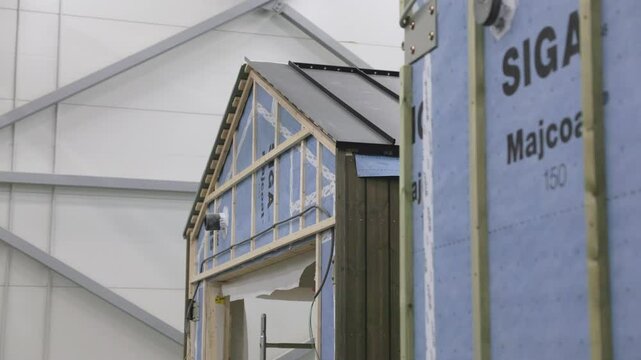 A partially constructed building with visible timber framing, SIGA Majcoat vapor barrier, and electrical wiring. Roofing and wooden cladding are being installed in an indoor workshop