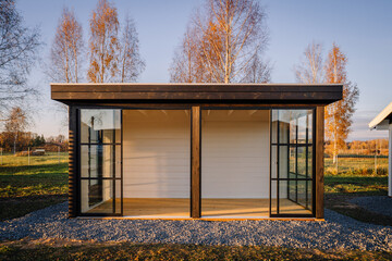 Modern black-framed glass garden house with sliding doors, surrounded by autumn trees, bathed in...