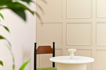 Minimalist interior with white round table, small decorative vase, wooden chair with green cushion, and beige paneled wall in background