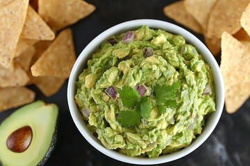 Obraz premium Authentic guacamole served in a white bowl surrounded by tortilla chips and a fresh avocado half, showcasing a healthy and delicious Mexican appetizer on a dark background.