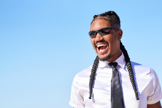 Stylish androgynous man smiling with braids and sunglasses against blue sky