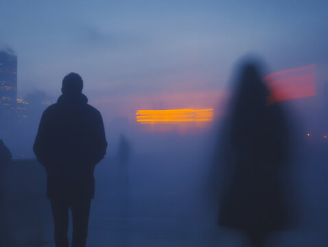 a photograph of two people standing at a distance, with their bodies turned away from each other, against a blurred cityscape background,