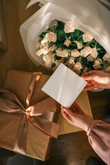 Hands holding a blank greeting card near a bouquet of delicate roses and a wrapped gift with a...