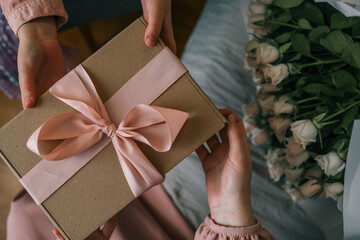 Hands of a child giving a wrapped gift with a satin ribbon to a woman holding a bouquet of roses,...
