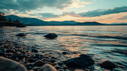 Obraz premium A serene lakeside view with rocks and gentle waves under a cloudy sky at dusk with mountains afar