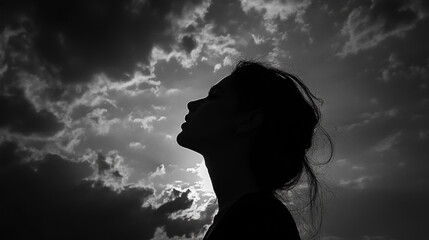 Silhouette of a woman looking up at the cloudy sky in black and white with bright light shining through