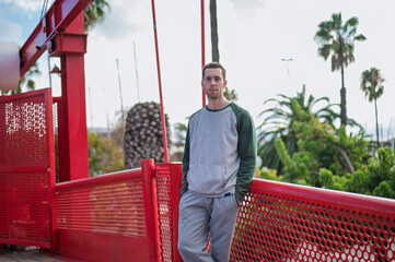 A young, blond-haired man in a gray tracksuit poses casually on a striking red bridge, smiling subtly, with palm trees and a bright sky in the background.