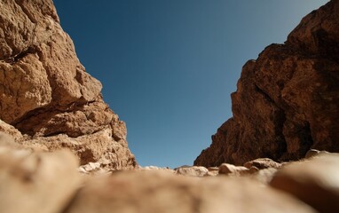 Fototapeta premium Low angle view of a rocky canyon under a vibrant blue sky. The canyon walls are tan and textured, with a shallow rocky path visible in the foreground. The image evokes a sense of vastness and