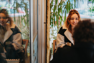 Two women enjoying a warm conversation in a cafe setting, with plants visible outside and cozy vibes. A reflective glass wall adds depth, capturing interactions and forming a layered composition.
