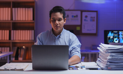 Young businessman working late in the office using laptop computer with graphs and charts showing on the screen and a large stack of paperwork on his desk