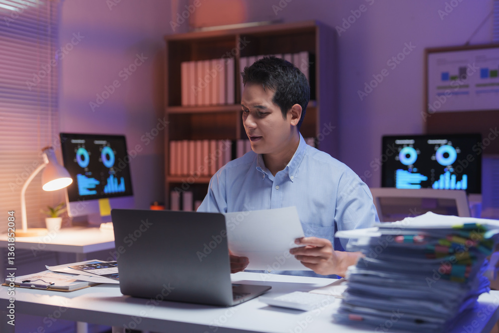 Poster young professional working overtime at night in office reading documents and using laptop, with grap