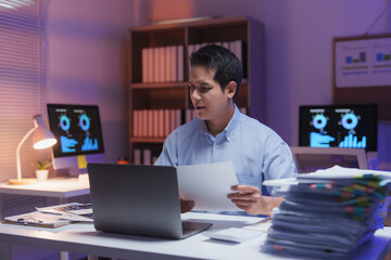 Young professional working overtime at night in office reading documents and using laptop, with graphs showing on computer screens, focused on work
