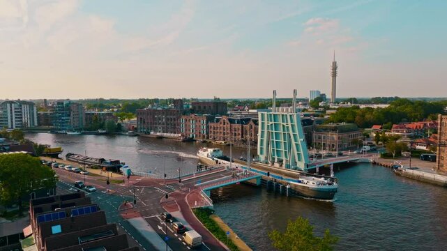 A cargo ship sails through a large draw bridge on a summer evening in Wormerveer, Netherlands. Historic warehouses, a river, and infrastructure are visible during golden hour. Captured in 4K drone