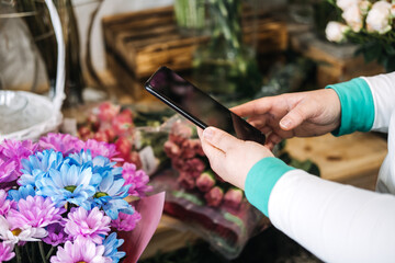 Florist using smartphone in flower shop to manage digital storefront. Reflects ecommerce floristry,...