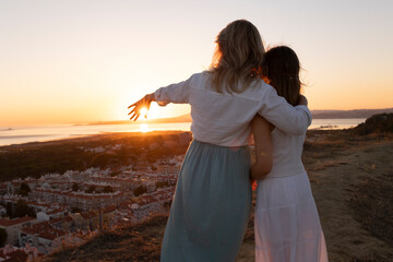 Fototapeta premium Mother and daughter embracing and pointing at sunset over coastal cityscape