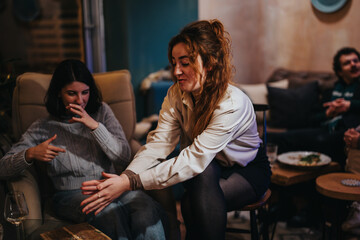 Two women share a casual, lively moment in a cozy lounge with friends enjoying food. The warm, inviting atmosphere reflects the joy of togetherness and a fun-filled gathering.