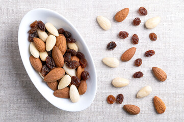 Almonds and raisins, a trail mix in an oval white bowl, on linen fabric. Also known as student food since 17th century, a mixture consisting of of almonds with seed coats and blanched, and of raisins.