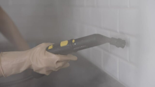Cleaning person uses steam cleaner on white tiles. Professional cleaner wearing protective gloves wielding steam cleaner, sanitizing white kitchen tiles with precision and thoroughness