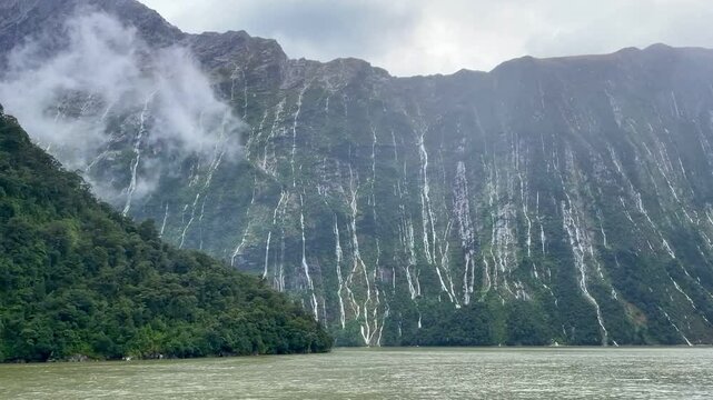 Scenic view of multitude of waterfalls falling from the dramatic mountain cliff during a heavy rainfall - Ferry Cruise Through Milford Sound fiord, Fiordland National Park, South Island, New Zealand