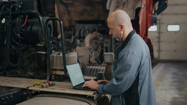 Side view medium shot of middle-aged Caucasian male mechanic working on laptop standing near semi-truck in professional auto repair shop
