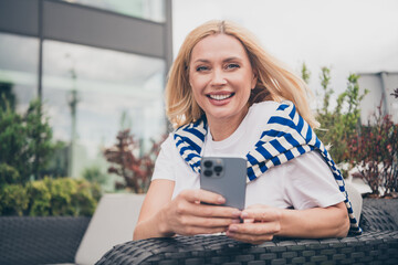 Cheerful middle-aged woman with blond hair using smartphone outdoors, enjoying sunny day