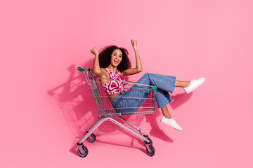 Happy young woman sitting playfully in a shopping cart against a pink background, radiating joy and...