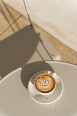A latte with leaf-shaped art sits on a table bathed in warm sunlight.
