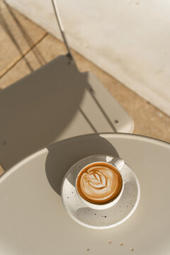 A latte with leaf-shaped art sits on a table bathed in warm sunlight.