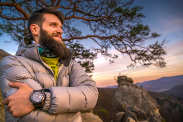 Bearded man in a puffer jacket stands on a mountain at sunset, arms crossed, wearing a Casio watch....
