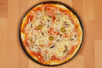 A top-down view of a baked pizza round pizza with ham, cheese, sliced mushrooms, and green olives on a wooden surface, ready to be served