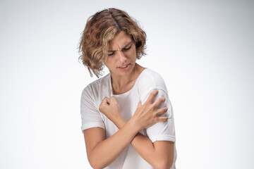 Caucasian woman in a white shirt gripping her shoulder tightly with a painful expression, showing...