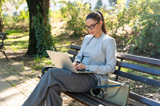 Successful smiling businesswoman in formal wear sitting on bench outdoor in park with plans using laptop computer working or having online meeting. Business, finance industry or real estate concept.
