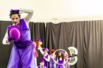Male clown wearing purple costume and holding purple bowler hat performing on stage with other blurred artists in background