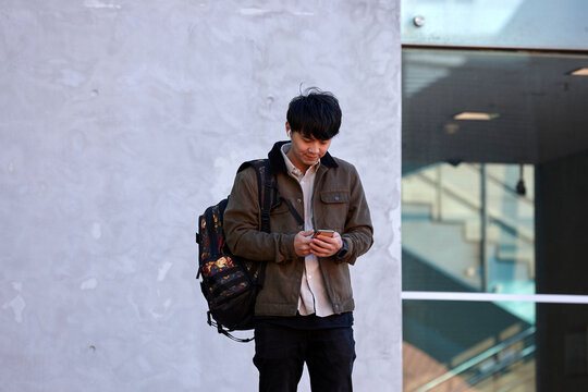 Young Chinese university student standing using mobile phone and wireless headphones