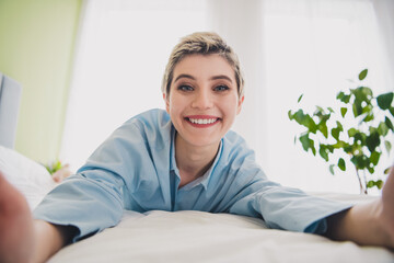 Young blonde woman in blue pajama smiling brightly while relaxing on a bed in a cozy, well-lit...