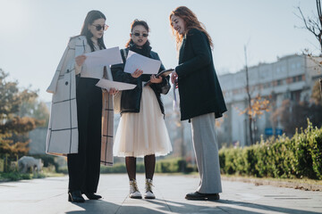 Three stylish women holding documents in an outdoor setting, discussing work-related strategies,...