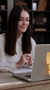 Young smiling brunette woman participates in an online interview and enthusiastically answers questions. Woman sits at an office table and talks in front of a computer explaining with her hands