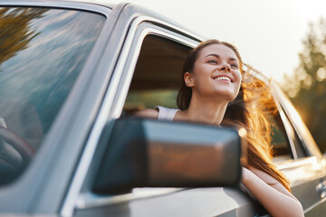Young woman enjoying a road trip, smiling with her hair flowing in the wind from the car window,...
